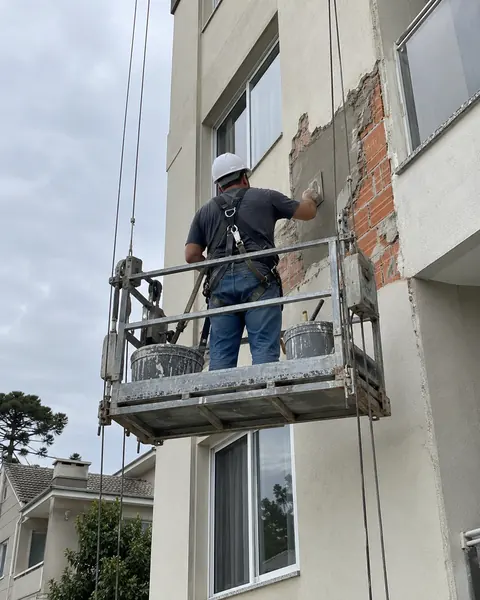 Trabalhador de capacete e cinto de segurança num balancim reparando o reboco da fachada de um prédio residencial